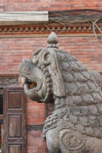 A close-up of a stone lion statue with intricate carvings, positioned against a backdrop of red brick wall and wooden window shutters.
