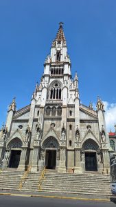 A tall, ornate church building with a pointed tower topped by a cross stands against a clear blue sky. The facade features intricate Gothic-style architecture, with detailed sculptures and multiple arched windows. A set of broad steps leads up to the main entrance, flanked by two large doors. The structure displays a combination of light and dark stone with a weathered appearance.