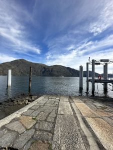 Stone boat ramp leading into a lake with mountains, a distant boat, and a "CANNOBIO" sign on a dock. The embarkation pier in Cannobio, a town located on the shores of Lake Maggiore in Italy.