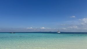A wide tropical ocean with clear turquoise water under a bright blue sky. A small dark boat and a white catamaran float in the distance, with a thin line of clouds on the horizon.
