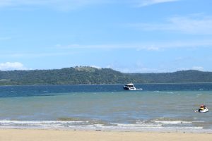 Sandy beach with gentle ocean waves, a motorboat and jet ski in the water, distant forested hills under a bright blue sky with scattered clouds.
