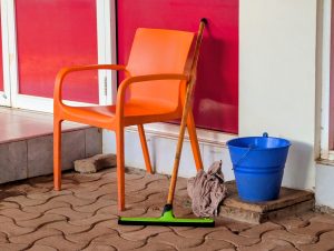 An orange plastic chair is positioned beside a blue bucket and a mop leaning against it. A gray towel is placed on the ground near the chair, which sits on a patterned stone floor.