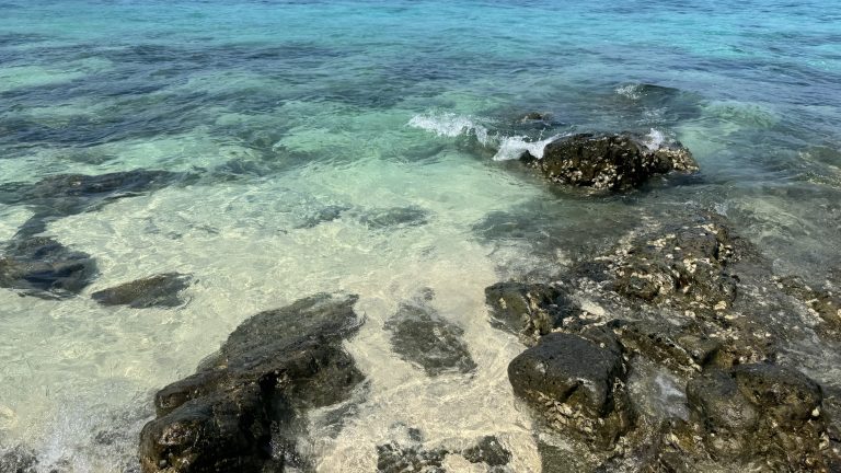 Clear turquoise water flows over dark rocks near the shore. You can see white sand and small stones beneath, with gentle ripples and tiny bubbles on the surface.