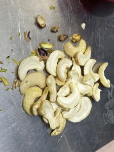 A pile of cashews and scattered pistachios on a metallic surface. The nuts are mostly whole, conveying a sense of snacking or food preparation.