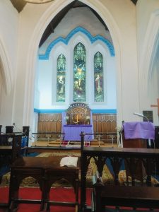 Church interior with a purple-draped altar, golden cross, and three stained glass windows above.