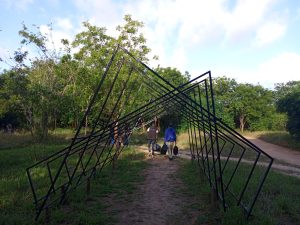Many people walk along a dirt path under a large triangular metal structure. The area is lush with trees and grass under a partly cloudy sky. The structure forms a striking canopy over the pathway.