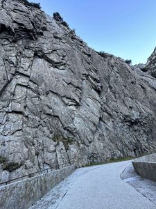 A winding path made of light gravel runs alongside a large, rocky cliff face. The cliff is composed of angular, weathered stone slabs, some covered with sparse vegetation at the top. The sky above is clear with a light blue hue, suggesting a serene outdoor environment.
