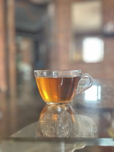 A clear glass cup filled with amber-colored tea sits on a reflective glass table, with a softly blurred indoor background.