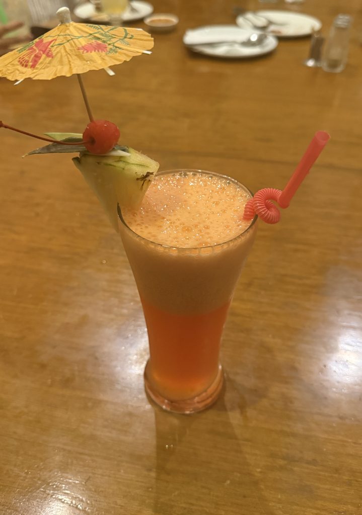 Tall, bubbly orange drink garnished with a cherry, pineapple slice, and paper umbrella on a wooden table.