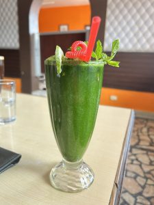 In the foreground a tall vivid green beverage in a clear glass with a red straw and curly garnish, topped with mint leaves and lime slice, sits on a light table. In the background white diamond patterned wall and an arch through to an orange wall area.Floor is crazy paving. 