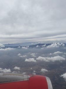 An aerial view from an airplane, showcasing a vast landscape below with rolling hills and patches of farmland. Fluffy white clouds are scattered throughout the scene, with a gray sky overhead. The airplane's wing is partially visible in the foreground, featuring a red color and metallic details. Mountains are faintly visible in the distance, adding depth to the horizon.