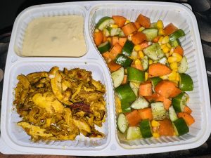 A white takeout container divided into three sections. One section contains seasoned, sliced, grilled chicken, golden brown and slightly crispy. The second section features a fresh salad made of diced cucumbers, carrots, and sweet corn, sprinkled with seasoning. The third section holds a small serving of creamy sauce, possibly a tahini or garlic sauce, with a smooth texture.