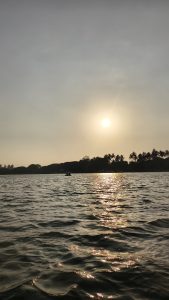 Sunset over a lake with golden light reflecting on rippling water. Silhouettes of trees and a small boat are visible against the evening sky.