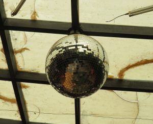 A classic mirrored disco ball hangs from a black metal frame against a weathered, translucent ceiling. Its numerous small glass facets catch and reflect the surrounding light, creating a shimmering effect.