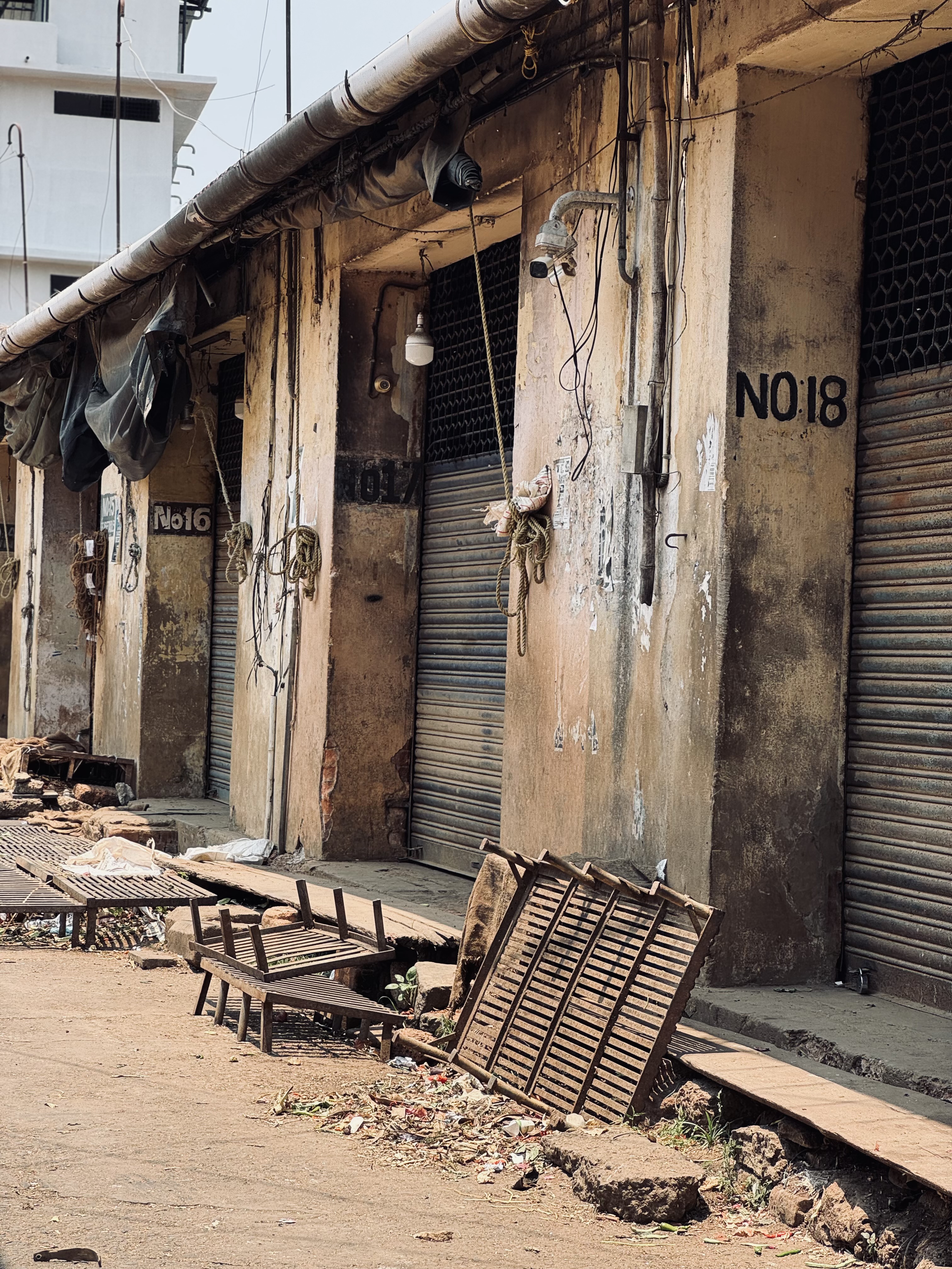 A rundown street featuring several closed shopfronts with rolling metal shutters at Palayam market. The walls of the shops are weathered and peeling, displaying numbers like "No 16," "No 17," and "No 18." Wires and fixtures hang loosely from the walls, and various debris, including wooden benches and scraps, are scattered across the ground