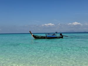A wooden longtail boat floats on calm, clear turquoise water under a deep blue sky with a few clouds.