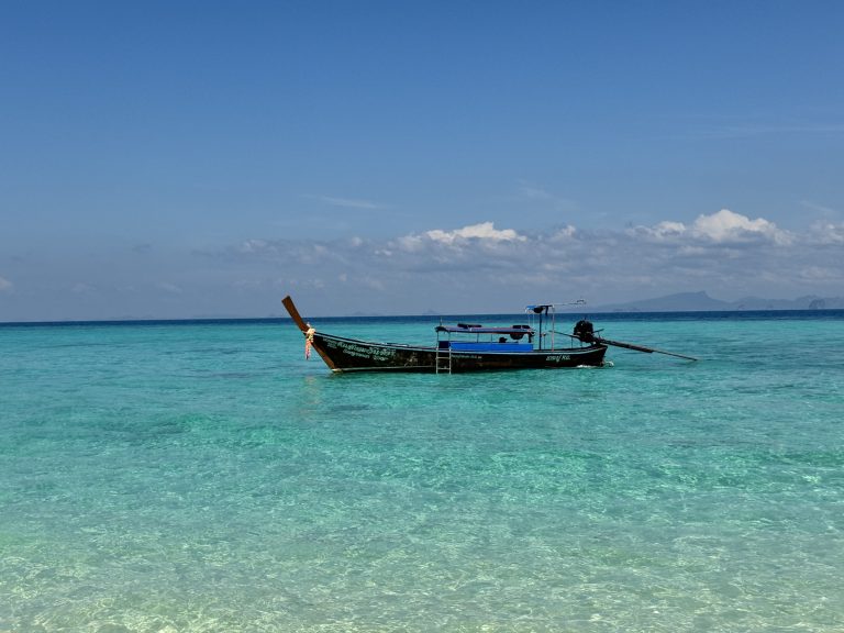 A wooden longtail boat floats on calm, clear turquoise water under a deep blue sky with a few clouds.