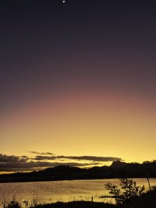 
A serene sunset over a calm lake, with hues of orange, purple, and blue blending in the sky. Silhouetted hills are visible in the background, and a crescent moon is faintly visible in the top left corner. 