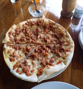 A thin-crust chicken pizza with melted cheese, sliced and served on a white plate with table condiments.