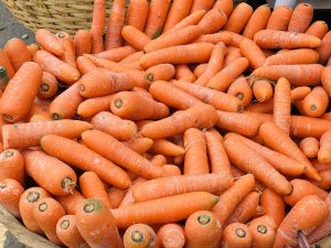 A large pile of fresh, bright orange carrots with varying lengths and sizes, arranged loosely among each other. In the background, a woven basket adds to the scene's rustic feel. 