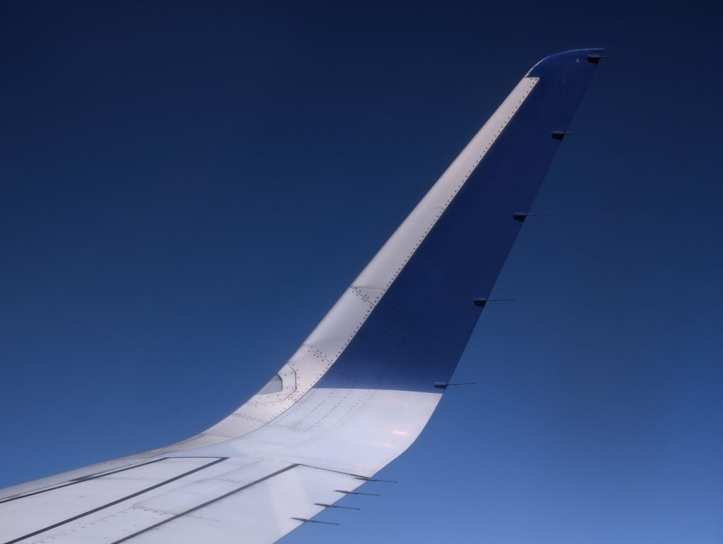 A close-up view of an airplane wingtip against a clear blue sky, highlighting the wing’s structure with its white and blue coloring, along with visible rivets and sensors.