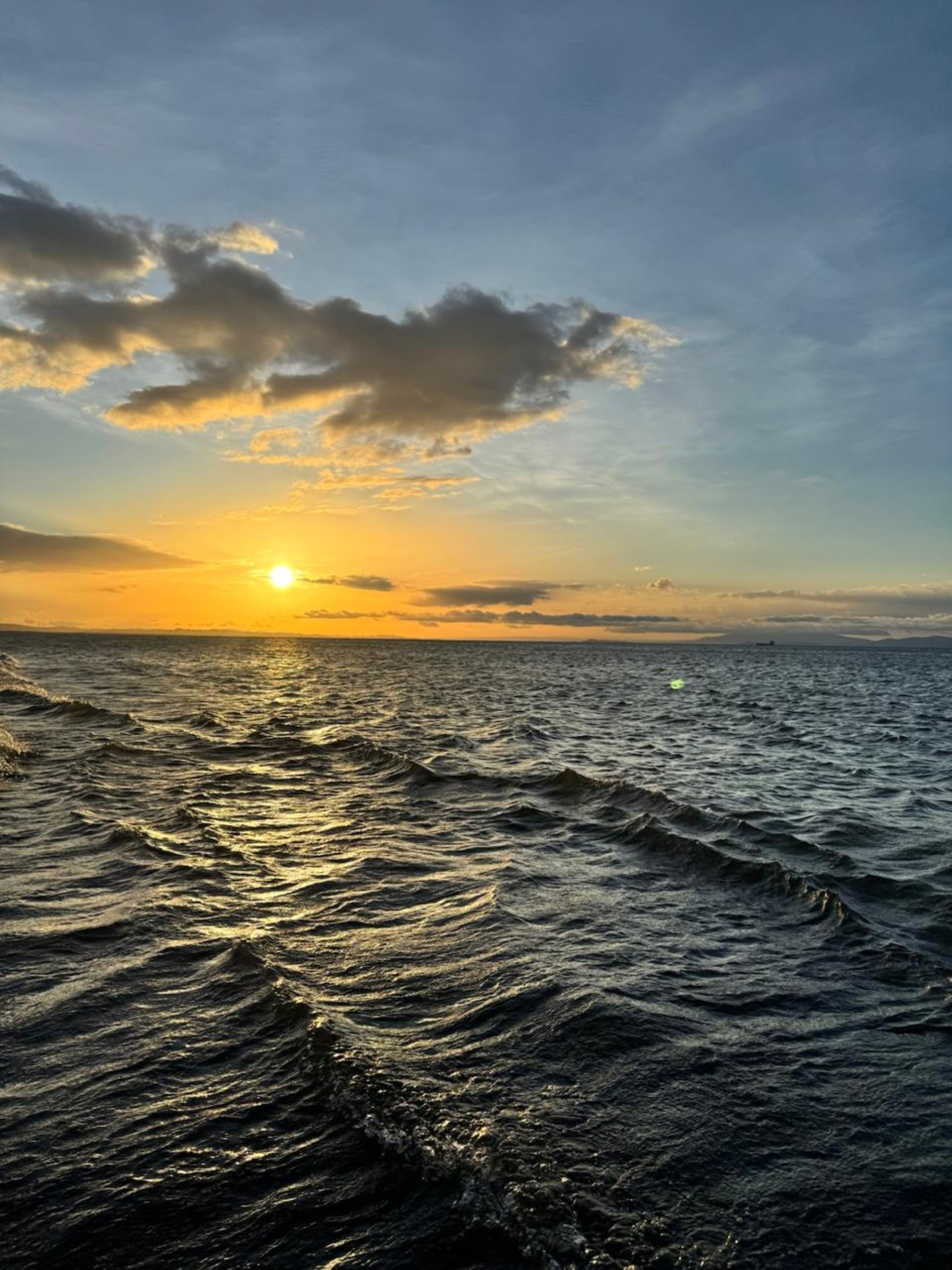 A serene seascape during sunset, featuring gentle waves reflecting golden light from the sun. The sky transitions from blue to orange and yellow hues as clouds drift across, adding depth to the tranquil scene.