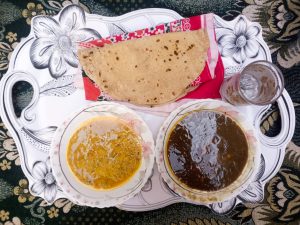 Top view of a traditional meal on a floral tray, featuring roti , a bowl of yellow lentil curry, a bowl of dark gravy curry, and a glass of water on a patterned cloth background.
