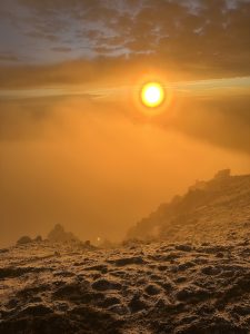 Sunrise from a high mountain peak, with orange light spreading over a sea of clouds and lighting up the rocky volcanic ground, creating a dramatic and beautiful view.
