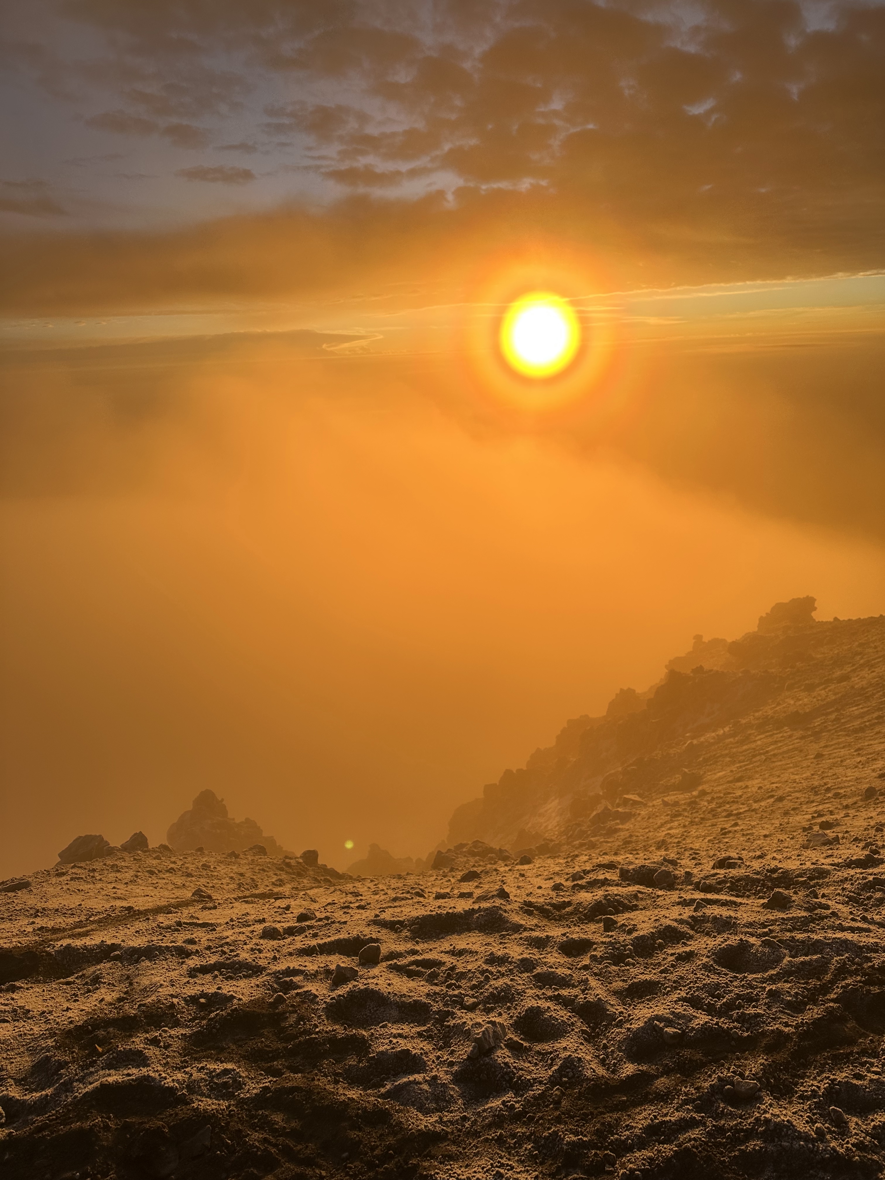 Sunrise from a high mountain peak, with orange light spreading over a sea of clouds and lighting up the rocky volcanic ground, creating a dramatic and beautiful view.