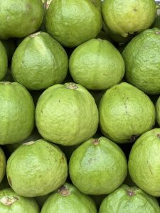 A close-up view of several glossy green fruits stacked closely together, showcasing their smooth and slightly bumpy surface. The fruits have a round shape with occasional small blemishes and stems visible on some of them, creating a vibrant and fresh appearance.
