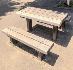 A wooden picnic table with attached benches sits on a paved outdoor surface, well-lit by the bright sun. The rectangular table and simple plank benches are supported by sturdy legs, with small patches of grass bordering the paving.
