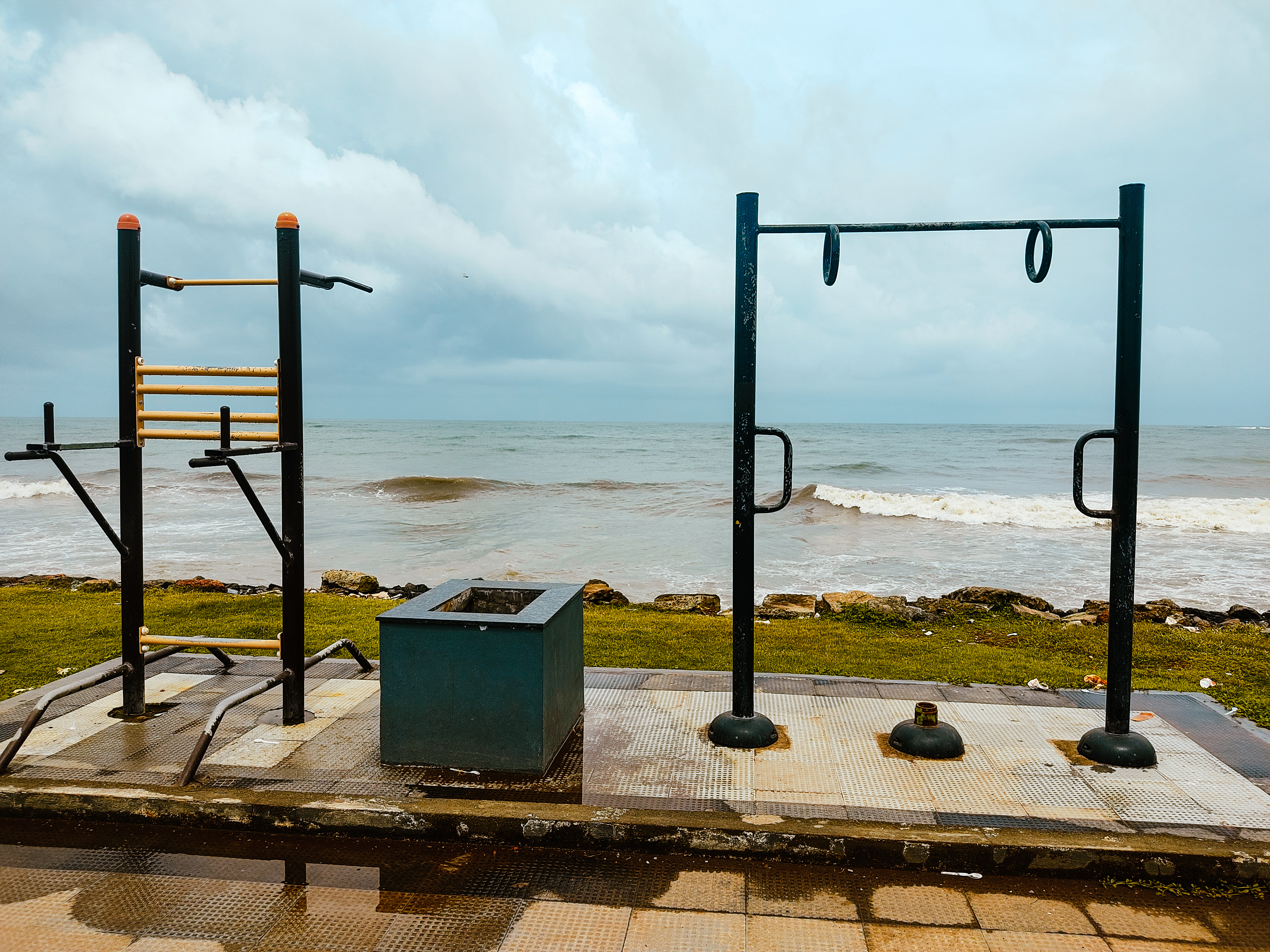 A beachfront workout area featuring outdoor exercise equipment, including a pull-up bar and a set of dip bars. The scene is set against a backdrop of the ocean with gentle waves, and the sky is overcast with gray clouds.