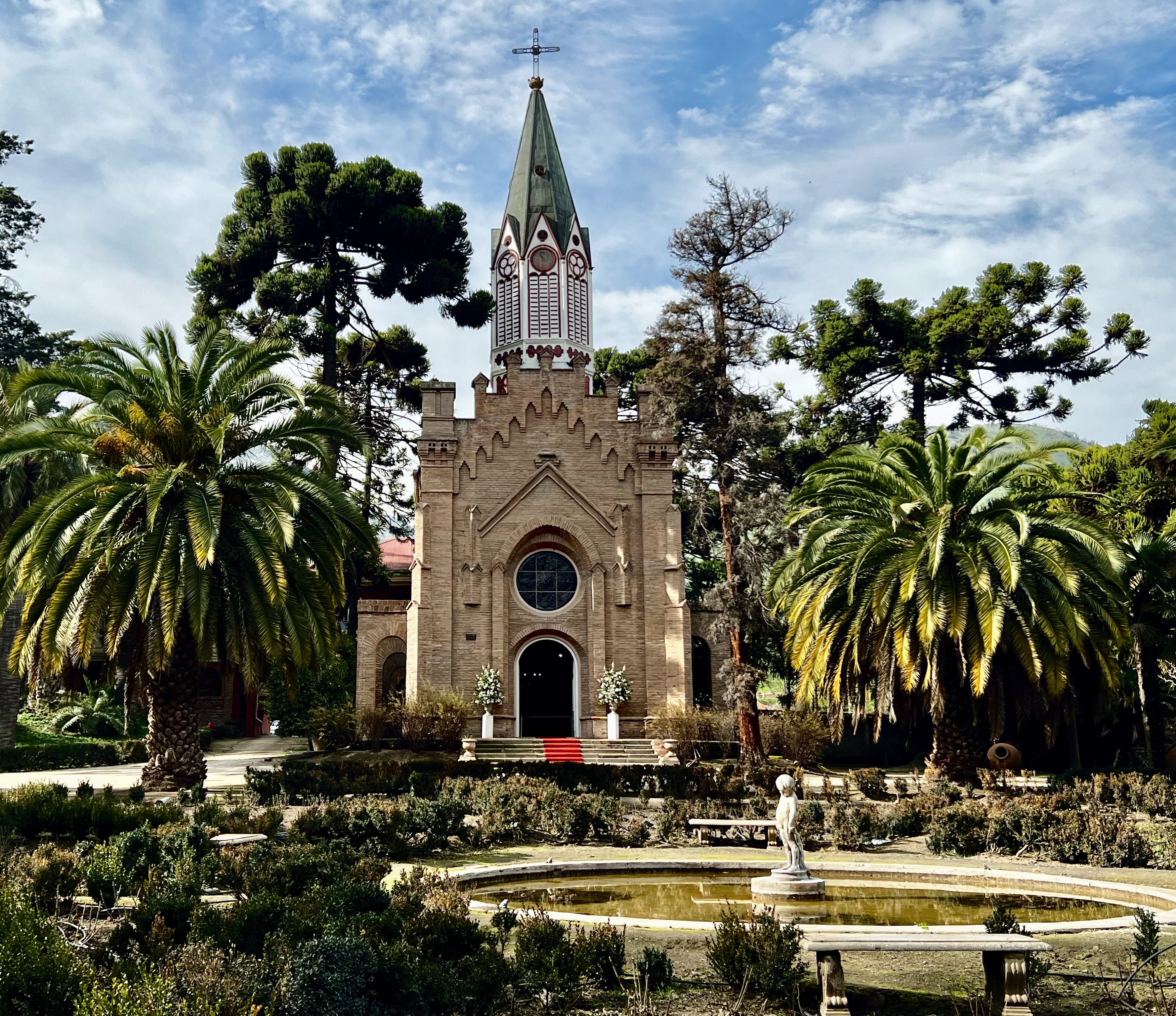A historic church with a green pointed roof, surrounded by palm trees and greenery in Santiago, Chile.