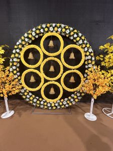 Circular floral backdrop made of yellow and white flowers with seven inner ring arrangements, each holding a hanging bell, set against a dark background. Decorative yellow flowering branches on stands are placed on both sides.

