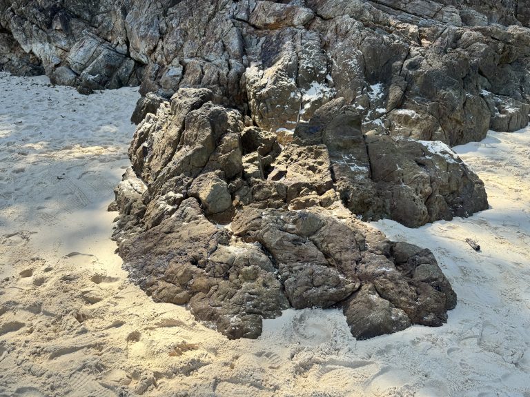 A large dark gray rock sits on a white sandy beach, rough and full of cracks. A rocky cliff rises behind it, and the sand is covered with footprints and small sunlit shadows.