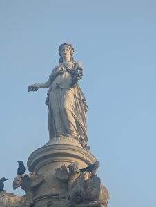 A tall, classical statue of a woman stands atop a weathered stone pedestal, holding a bouquet of flowers in one hand. 
