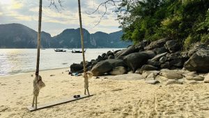 A simple wooden swing hangs from ropes on a sandy beach, with rocks, bushes, calm ocean, boats, and rocky islands under a soft evening sky.