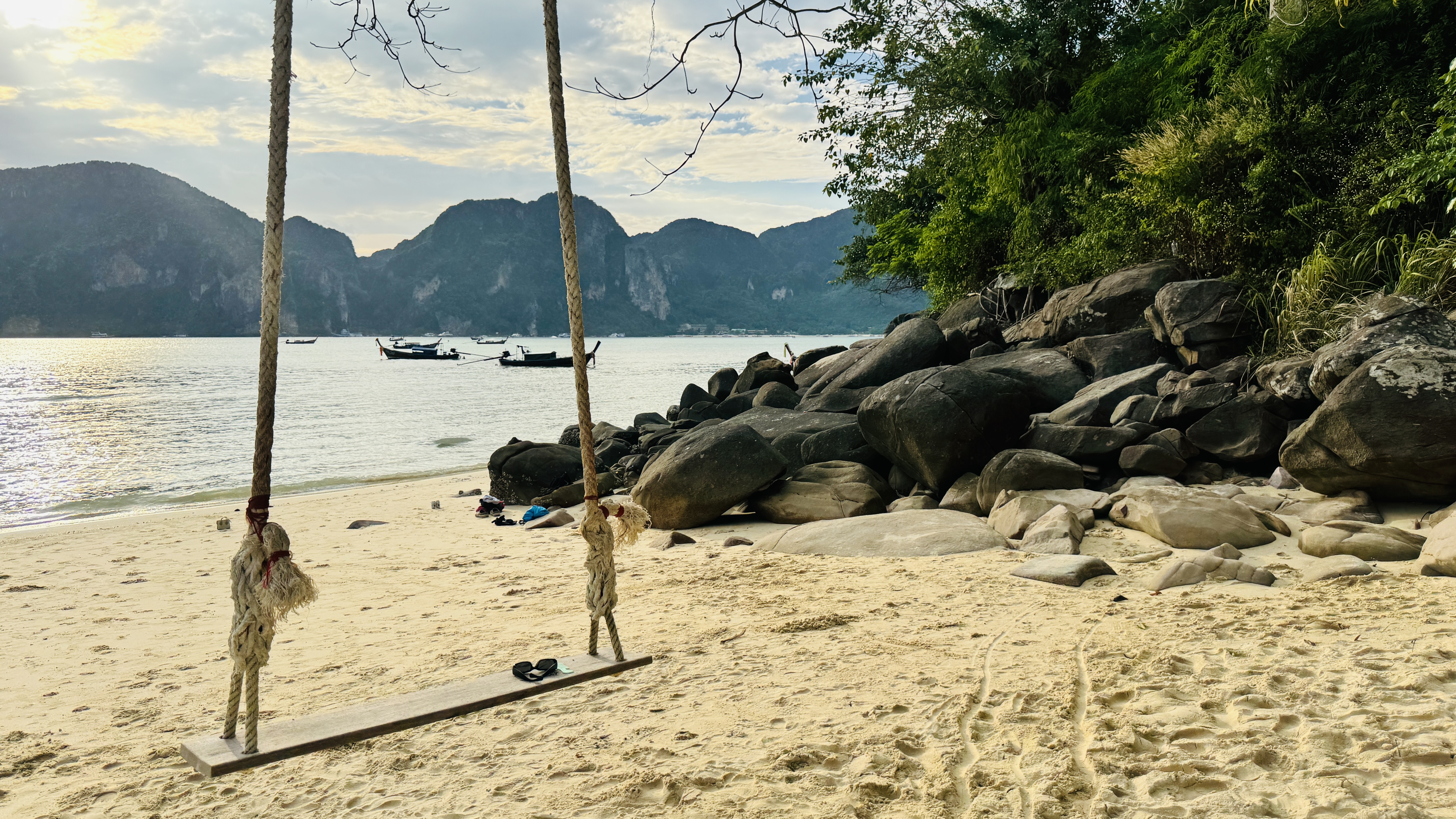 A simple wooden swing hangs from ropes on a sandy beach, with rocks, bushes, calm ocean, boats, and rocky islands under a soft evening sky.