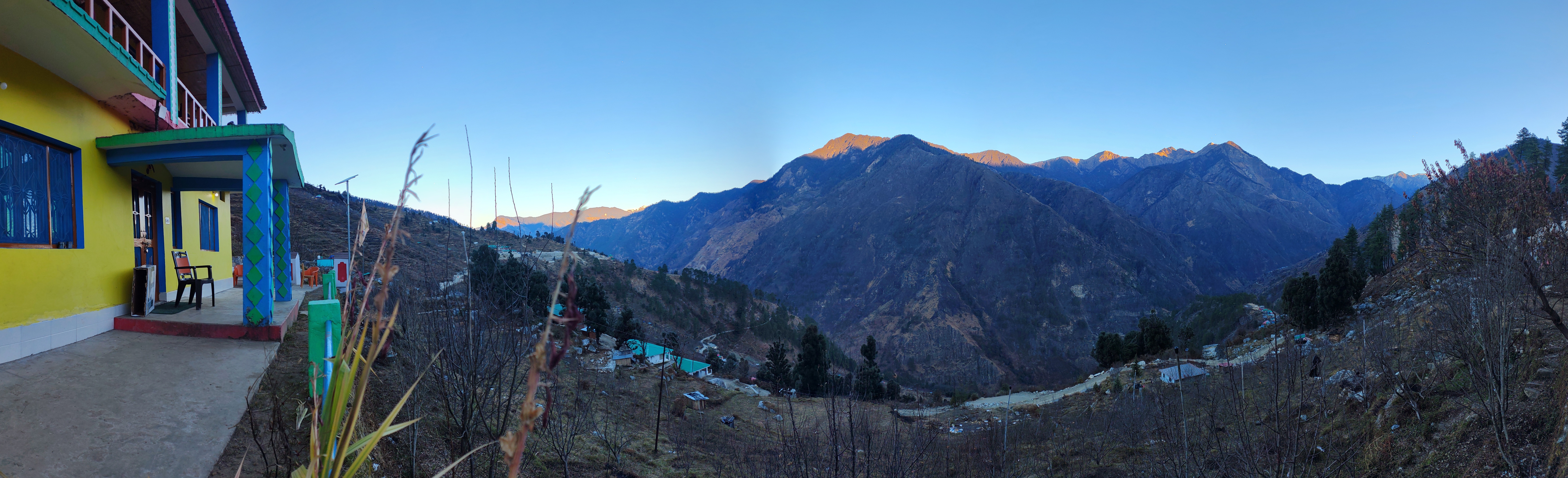 A sunrise view of a mountain village with a yellow house on the left, and a wide valley with hills and scattered homes under a clear blue sky.