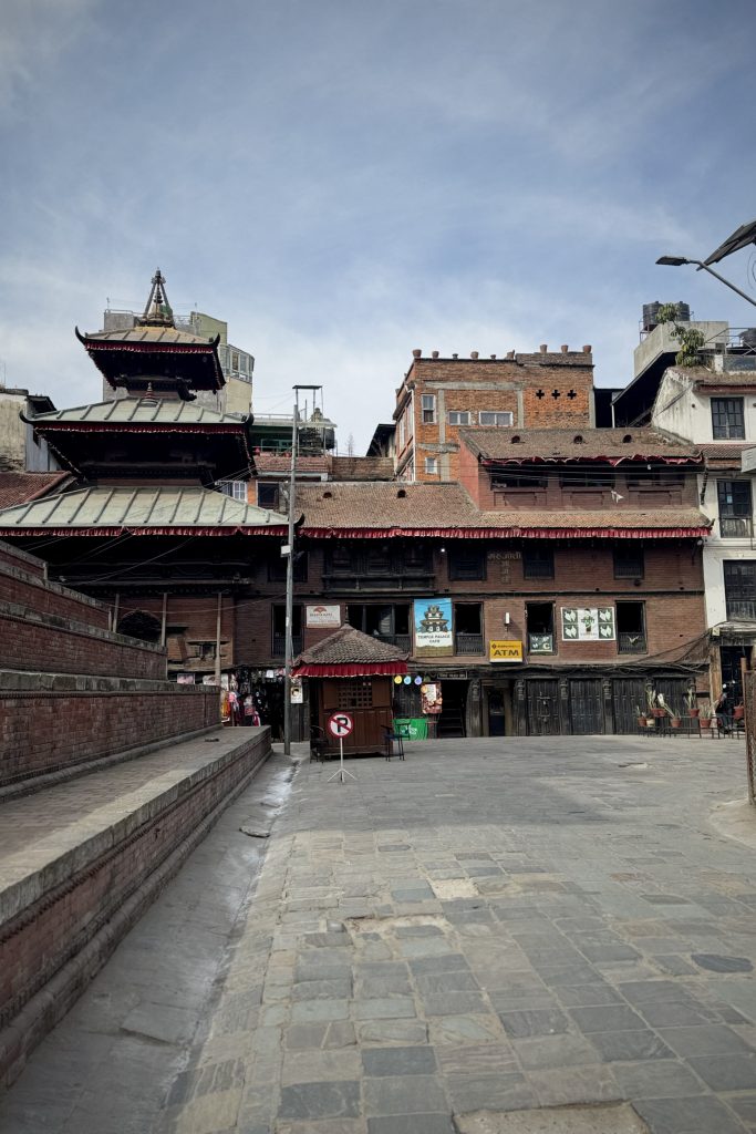 A narrow street in an urban area featuring an array of traditional and modern buildings. In the foreground, there is a stone-paved walkway with a low stone wall on the left. To the right, a small structure with a red awning stands in front of a larger building displaying various signs, including one for an ATM. Above, a multi-tiered temple-like structure with ornate roofing contrasts with the surrounding brick and concrete buildings. The sky is partially cloudy, giving a moody ambiance to the scene.