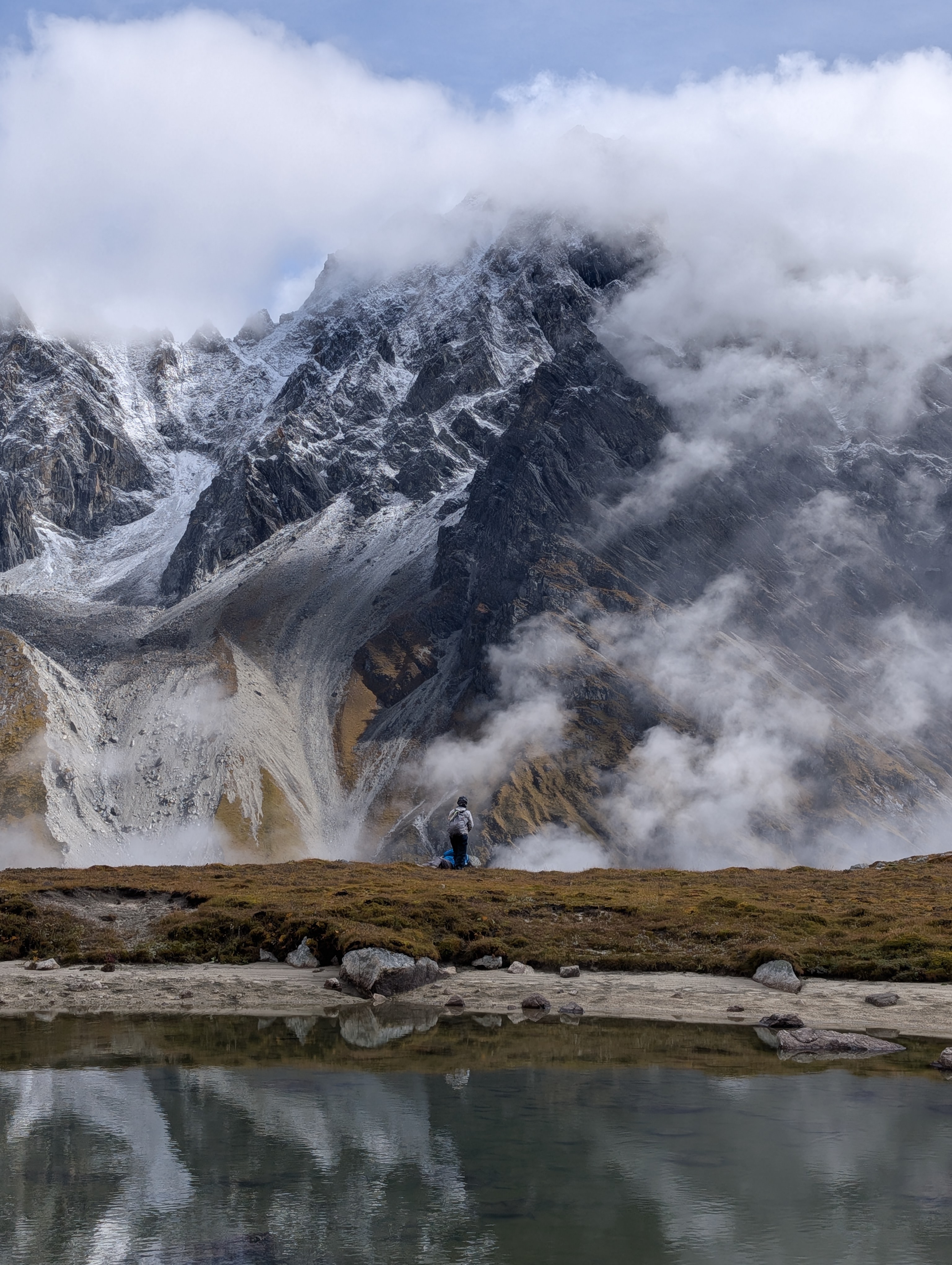 Snow-covered rocky mountain landscape with clouds partially covering distant peaks under a blue sky.