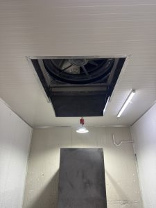 A view looking up at a metallic ceiling vent with a circular fan inside, set into a clean, light-colored ceiling. Below the vent, there is a large, flat, gray surface or panel, illuminated by a hanging light fixture. The walls of the room are smooth and light-colored, and there is a white pipe running along one wall. The overall atmosphere appears industrial or functional.