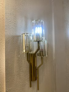 Gold wall sconce with three cylindrical glass shades, emitting a soft, warm glow. The textured background adds an elegant, cozy ambiance.