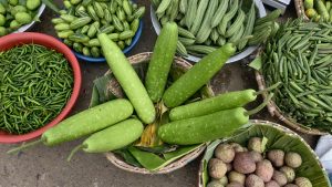 A top down view of a market stall featuring baskets of long green bottle gourds, okra, ridge gourd, and small pointed gourds alongside a bowl of green chili peppers.