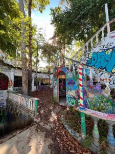 A partially overgrown area featuring an abandoned building with colorful graffiti on its walls. The structure includes an ornate staircase with a balustrade, leading to various openings, and surrounded by trees and fallen leaves. Sunlight filters through the trees, casting shadows on the ground, enhancing the vibrant colors of the graffiti and the natural surroundings.
