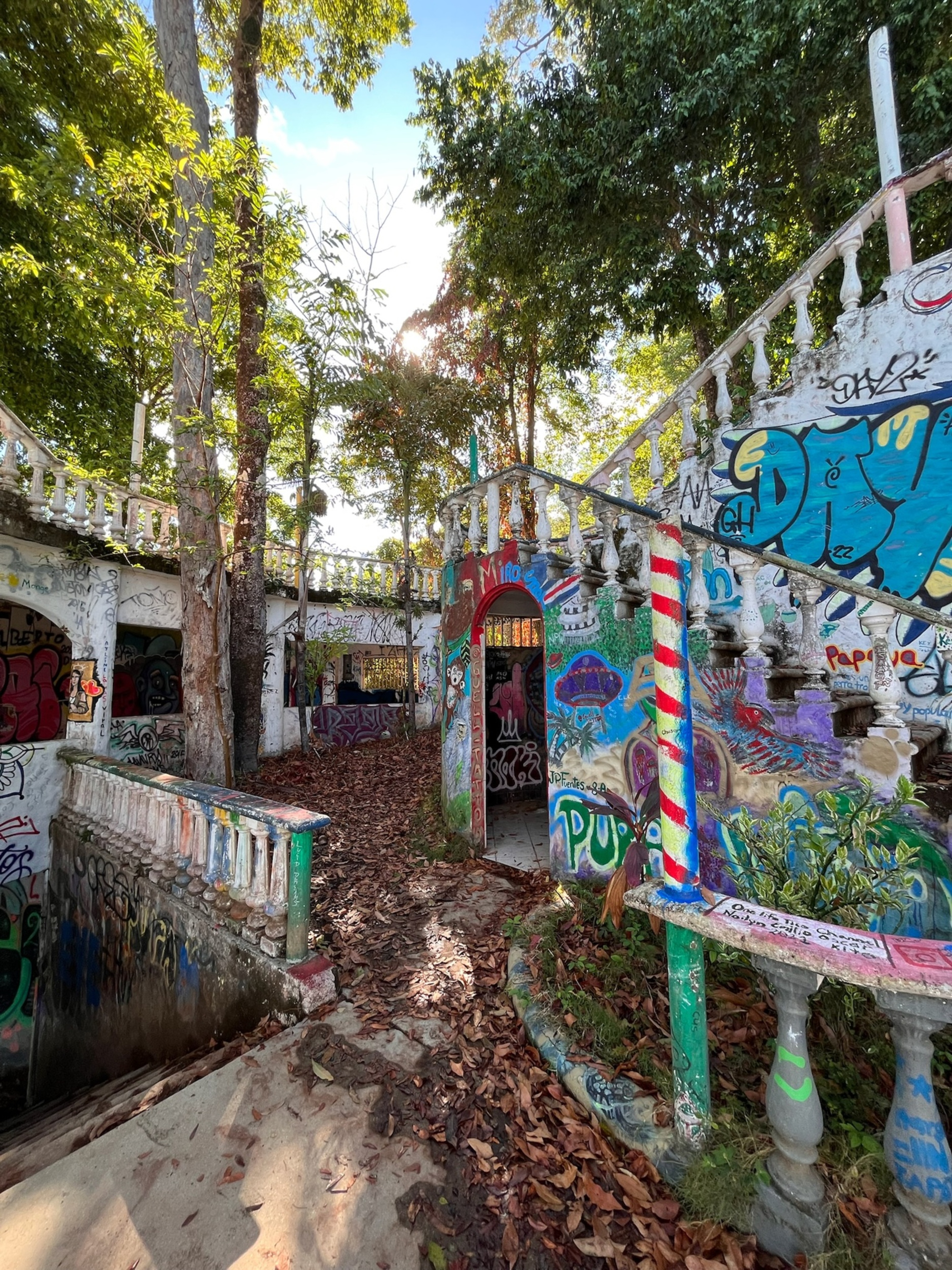 A partially overgrown area featuring an abandoned building with colorful graffiti on its walls. The structure includes an ornate staircase with a balustrade, leading to various openings, and surrounded by trees and fallen leaves. Sunlight filters through the trees, casting shadows on the ground, enhancing the vibrant colors of the graffiti and the natural surroundings.