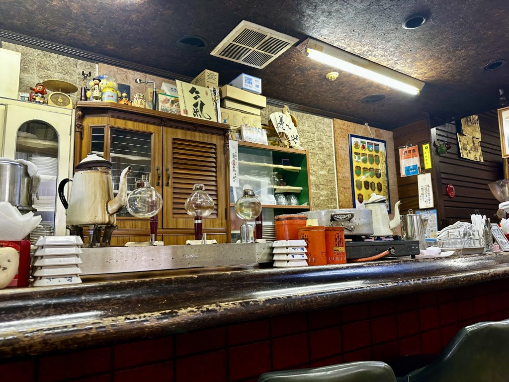 The worn wooden counter of an old Japanese kissaten (coffee shop) in Yonezawa, Yamagata, holds vintage siphon coffee brewers, a Sanko Coffee tin, and stacked dishware. A louvred wooden cabinet, Japanese calligraphy signs, a paper fan, and decades of accumulated memorabilia cover the walls behind.