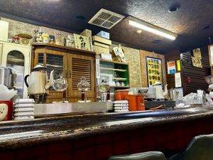 The worn wooden counter of an old Japanese kissaten (coffee shop) in Yonezawa, Yamagata, holds vintage siphon coffee brewers, a Sanko Coffee tin, and stacked dishware. A louvred wooden cabinet, Japanese calligraphy signs, a paper fan, and decades of accumulated memorabilia cover the walls behind.