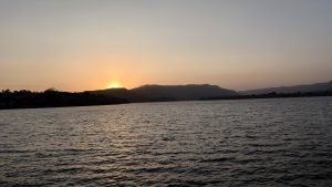 A serene sunset over a tranquil body of water, with gentle ripples reflecting warm hues of orange and yellow. Silhouetted hills rise in the background, creating a peaceful landscape that captures the beauty of dusk.
