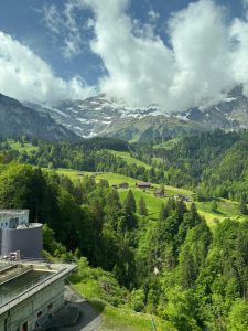 This image shows a beautiful mountain landscape with lush green hills and dense forests in the foreground. In the middle of the scene, small houses are scattered across the hills, creating a peaceful rural setting. In the background, tall mountains rise with patches of snow on their peaks, partially covered by large, dramatic clouds.